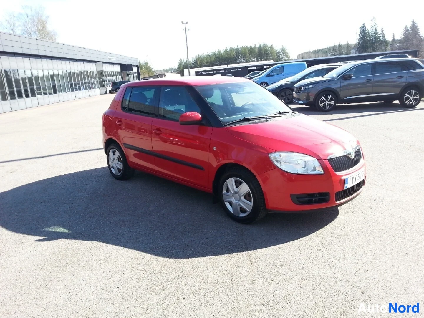 Etuviisto, nokka oikealle: Punainen viisiovinen Skoda Fabia -henkilöauto kuvattu etukulmasta oikealta, pysäköitynä auringonpaisteessa autotallin pihalla.