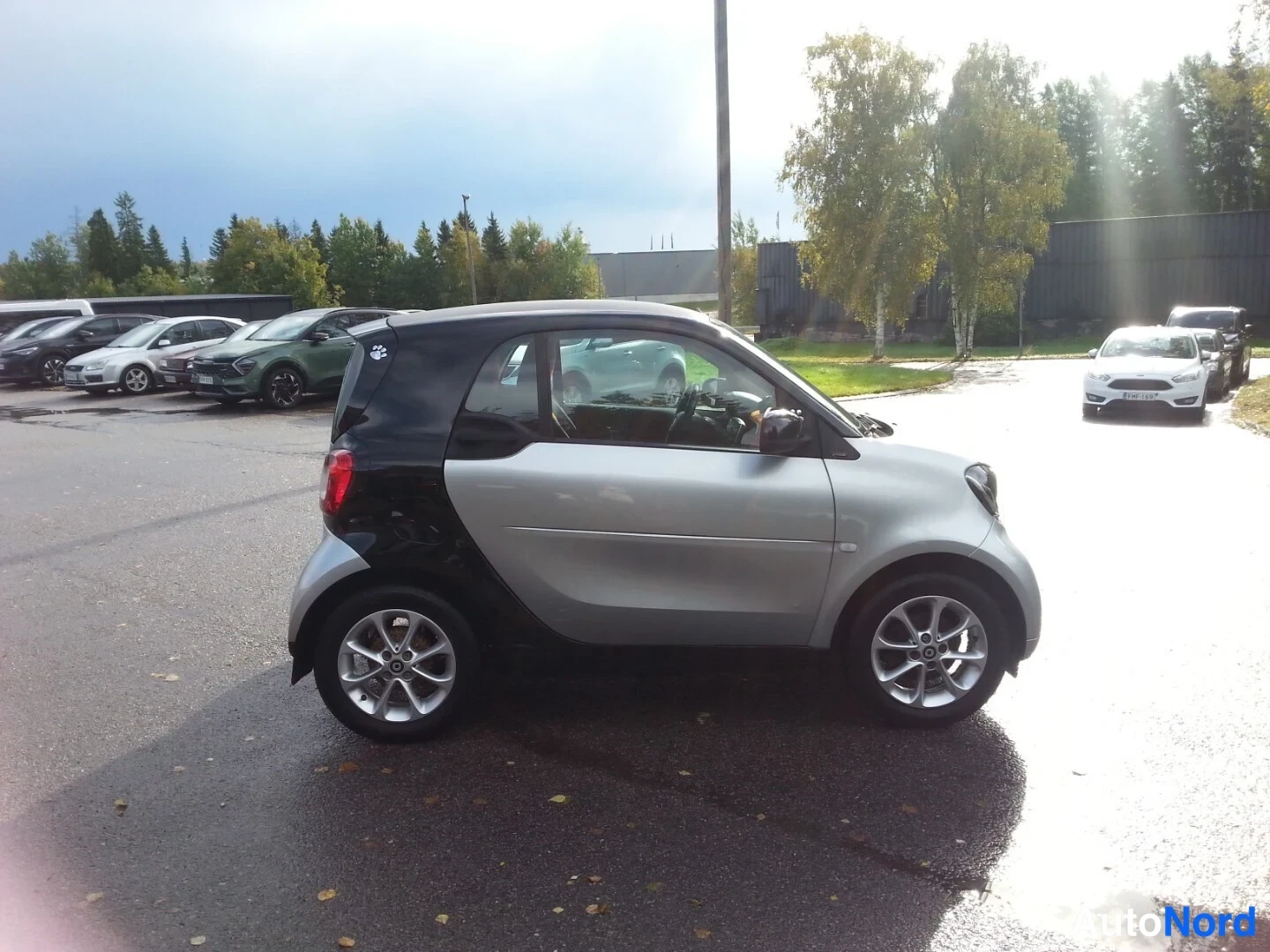 Etuviisto, nokka oikealle: Hopeanvärinen Smart Fortwo kolmannes-etuvasemmalle kuvattuna, tumma katto ja eturunko, hopeaiset ovipaneelit ja kevytmetallivanteet, pysäköintialueella.