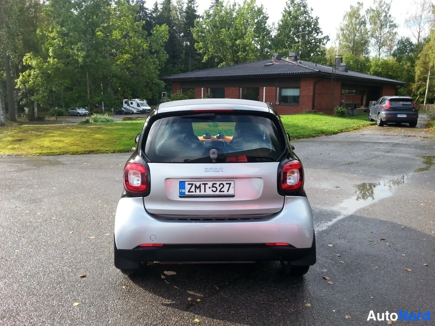 Takaa: Auton takaosan suora kuva: hopeanvärinen Smart, rekisteritunnus, punaiset takavalot ja tumma katto, parkkipaikka märällä asfaltilla.