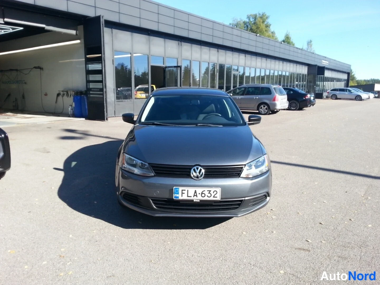 Edestä: Volkswagen Jetta edestäpäin harmaana, rekisterikilpi, paikoillaan autoliikkeen edustalla.