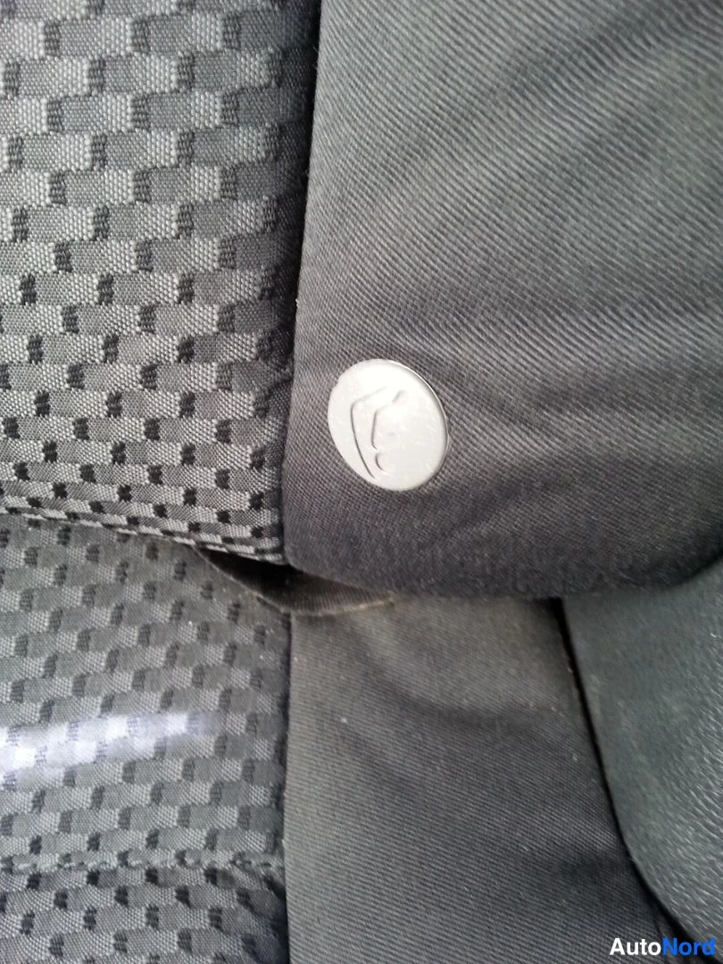 Yksityiskohta: Sisäkuva harmaasta kankaanverhoisesta autonistuimesta, jonka sivussa on pyöreä metallinen embleemi.