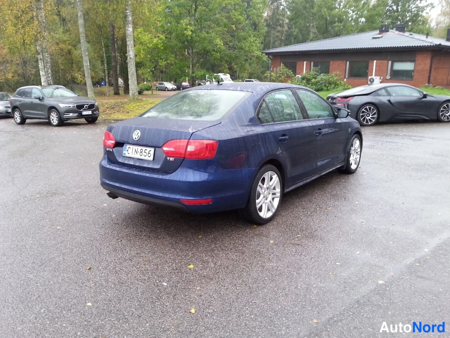 Takaviisto: Tummansininen Volkswagen Jetta -sedan takanurkka oikealta, takavalot ja rekisterikilpi IN-856 näkyvissä sateisella parkkipaikalla.