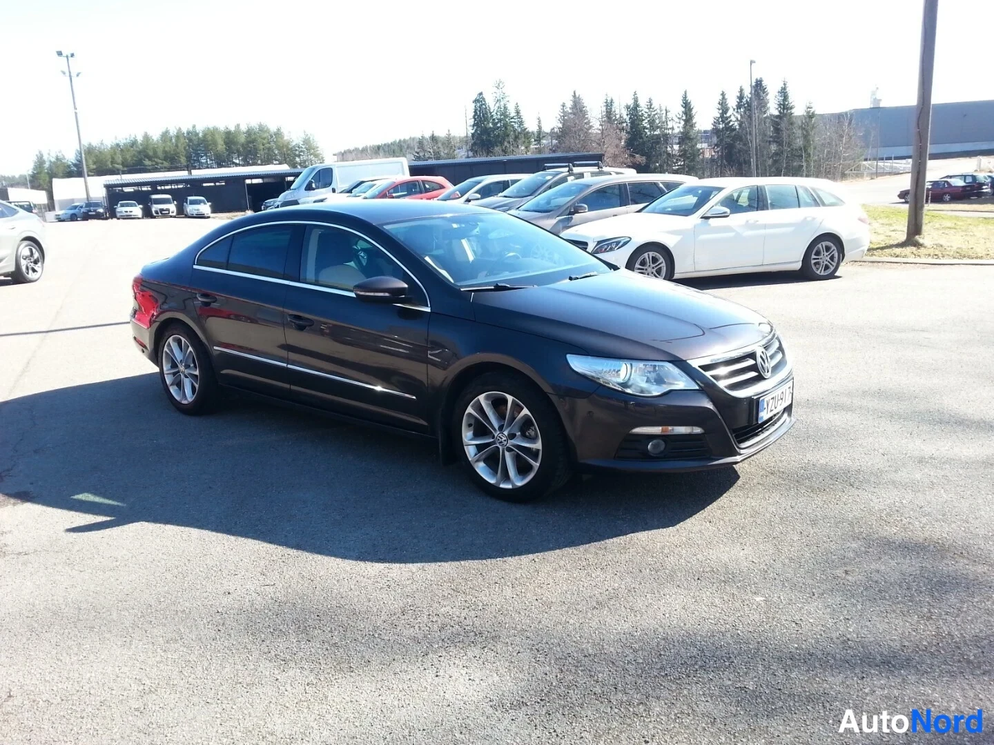 Etuviisto, nokka oikealle: Volkswagen Passat CC tummanruskeana etu kolmasosakulmasta, keula oikealla, valmis pingottu auringossa huoltoaseman pihalla.