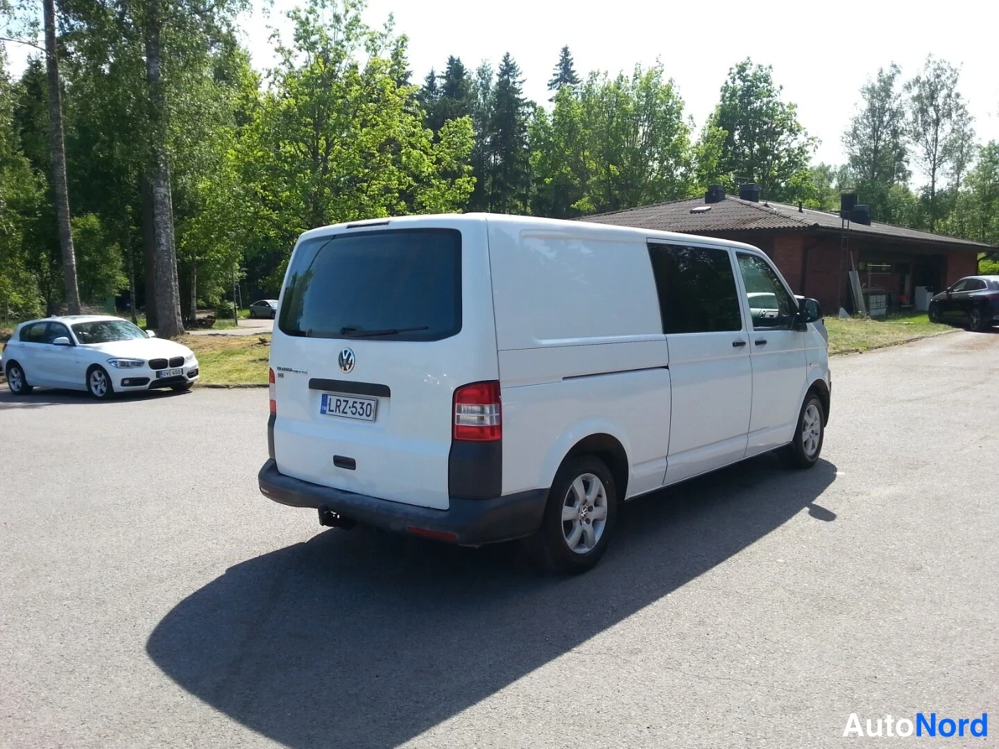 Takaviisto: Valkoinen pakettiauto takaviistöstä, rekisterikilpi ja lisävalaisin takapuskurissa, asfalttipihalla puiden ja punaisen rakennuksen taustalla.