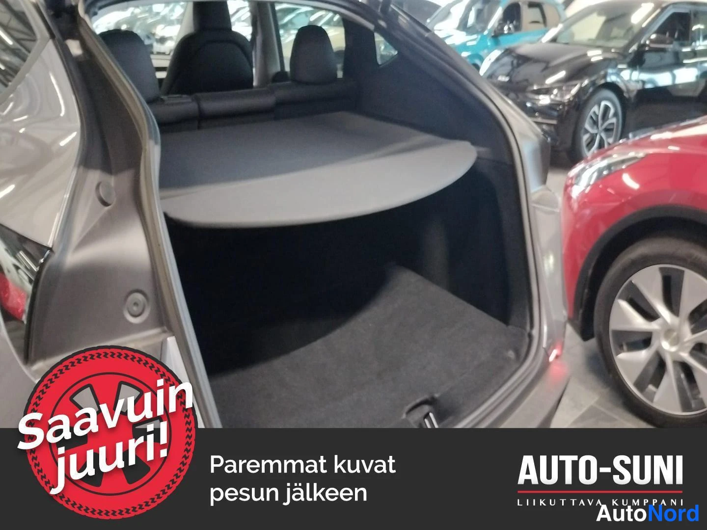 Muu: Auton avattu tavaratila, jossa tumma verhoilu, irtain takaselin suoja ja matonurkka; taustalla autosalin muita autoja.