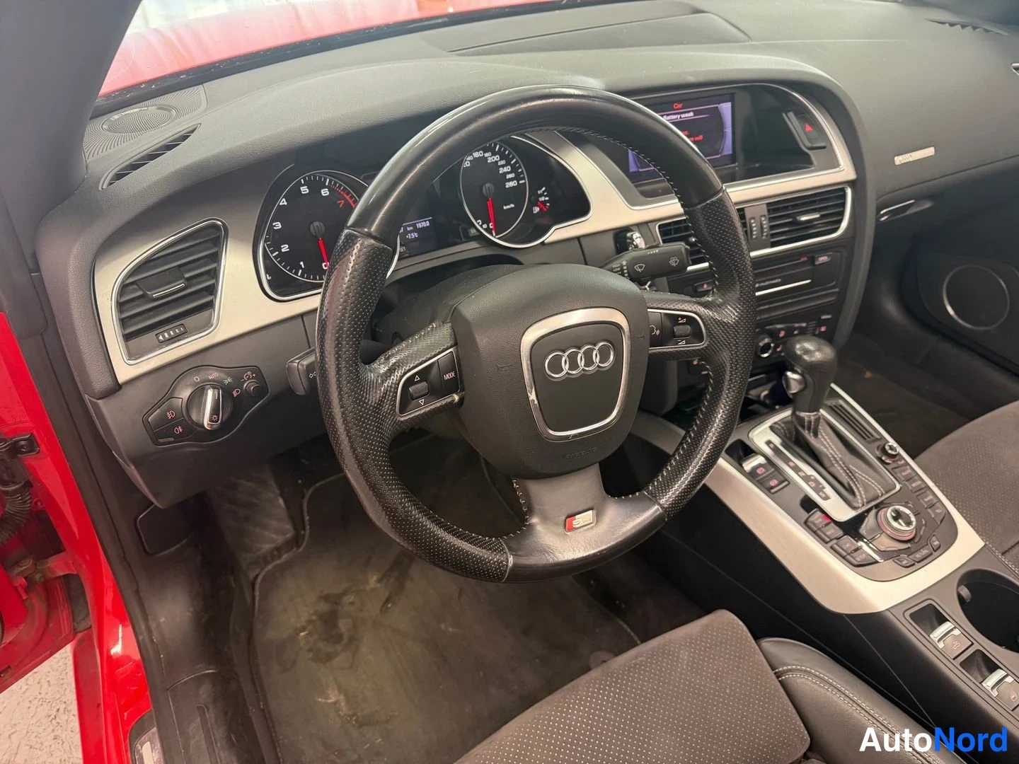 Sivulta: Auton etupenkin ohjaamoon kohdistettu lähikuva: ratin keskellä Audi-embleemi, mittaristo, radiopaneeli ja automaattivaihteiston vaihdekeila.