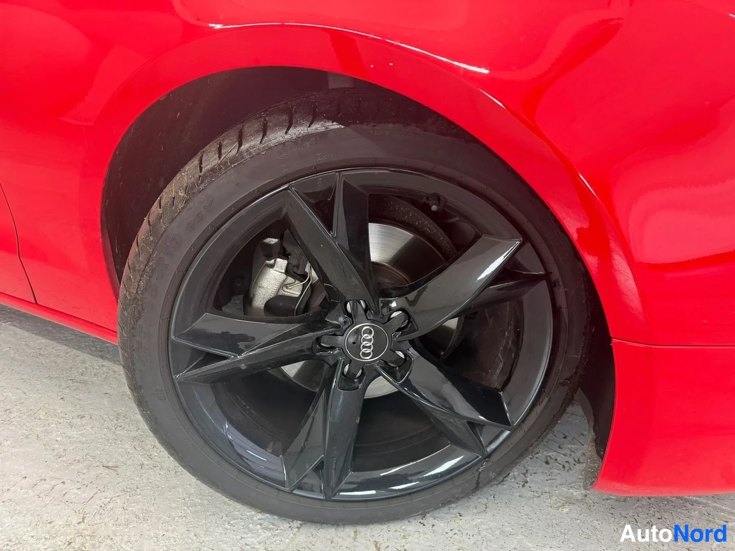 Sivulta: Punaisen auton pyörä lähikuvassa: musta vanteen pinnan kiiltävä pinta, Audi-merkki keskellä ja jarrusatula.