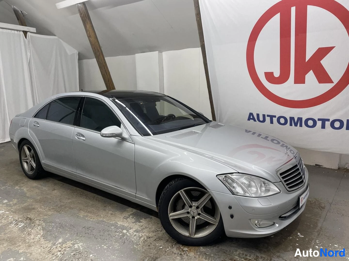 Etuviisto, nokka oikealle: Hopeanharmaa Mercedes-Benz -sedan etuosalta ja vasemmalta sivulta sisätiloissa, JK Automotor -tausta.