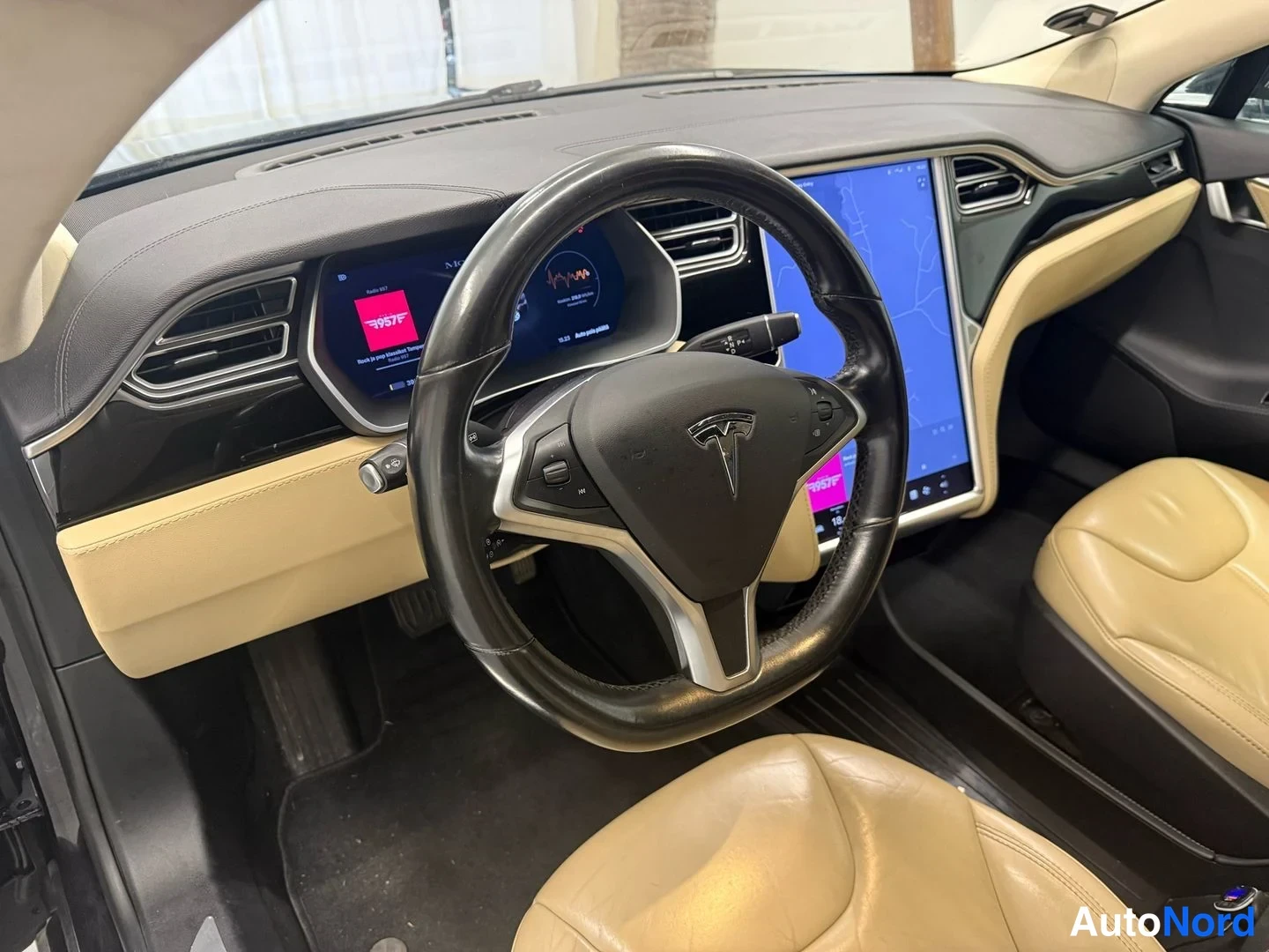 Muu: Tesla Model S:n sisätilat kuljettajan puolelta: musta ohjauspyörä, digitaalinen mittaritablo, pystysuora keskinäyttö sekä beigeä nahkaa penkissä ja ovissa.