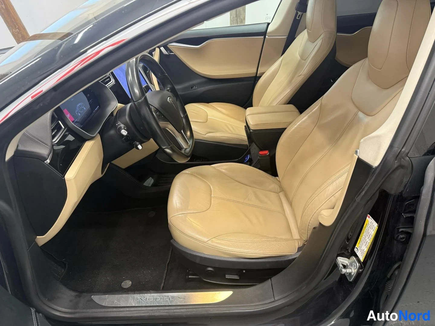 Muu: Tesla Model S:n etuistuinrata avatusta ovesta: beigeä nahkaa verhoillut etupenkit, musta ohjauspyörä, keskikonsolin ikkunaverho ja kynnyksellä Model S -teksti.
