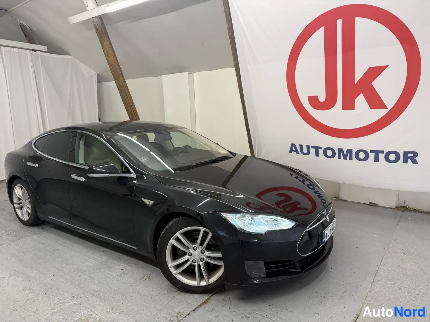 Etuviisto, nokka oikealle: Tesla Model S -sedanin kulmaotos edestä oikealta: musta koripinta, halo-kevytmetallivanteet ja rekisterikilpi näkyvissä sisätiloissa, taustalla Jk Automo