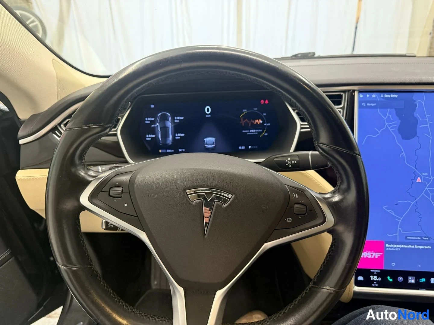 Muu: Tesla Model S:n kuljettajan näkymä: musta ohjauspyörä T-logolla, digitaalinen mittaritablo sekä keskikonsolin suuri kosketusnäyttö karttaan ja musiikkiin avattu