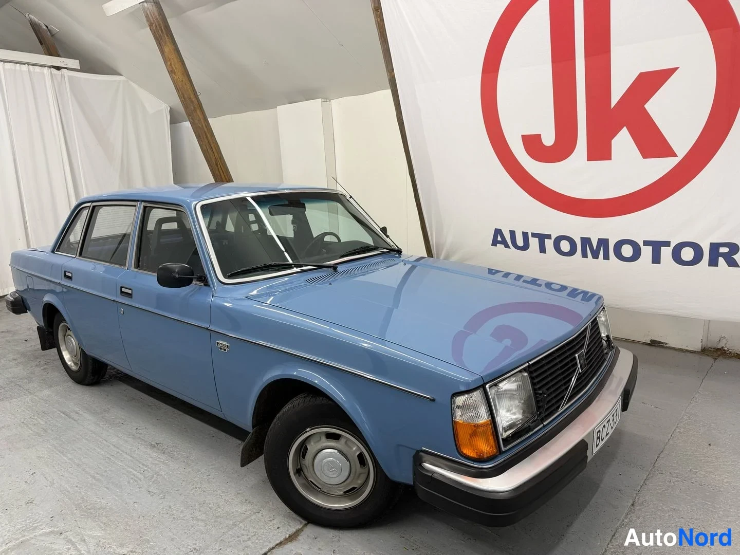 Etuviisto, nokka oikealle: Sininen Volvo 240 -sedan etukenänurkkakuvassa sisätiloissa; etupanssari, etupyörä ja kylki näkyvät.