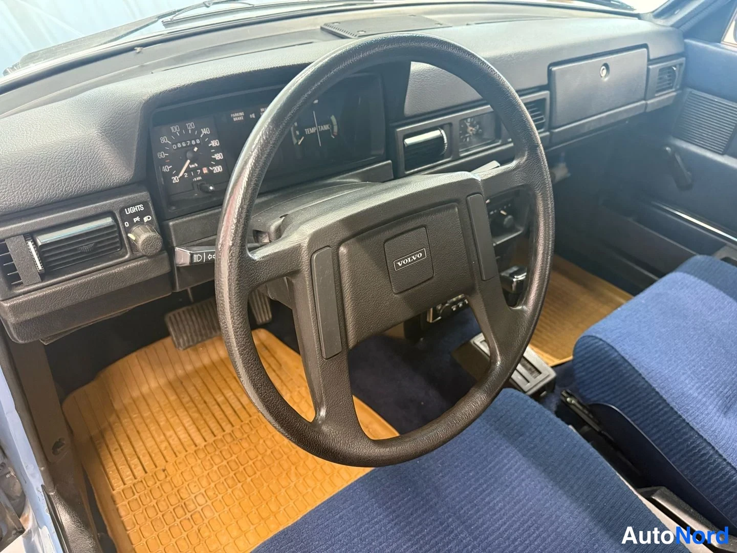 Muu: Ohjaamo näkyvästi: Volvo-merkkinen ohjauspyörä, mittaristo, tumma kojelauta, sininen istuin ja keltainen mattolattia.