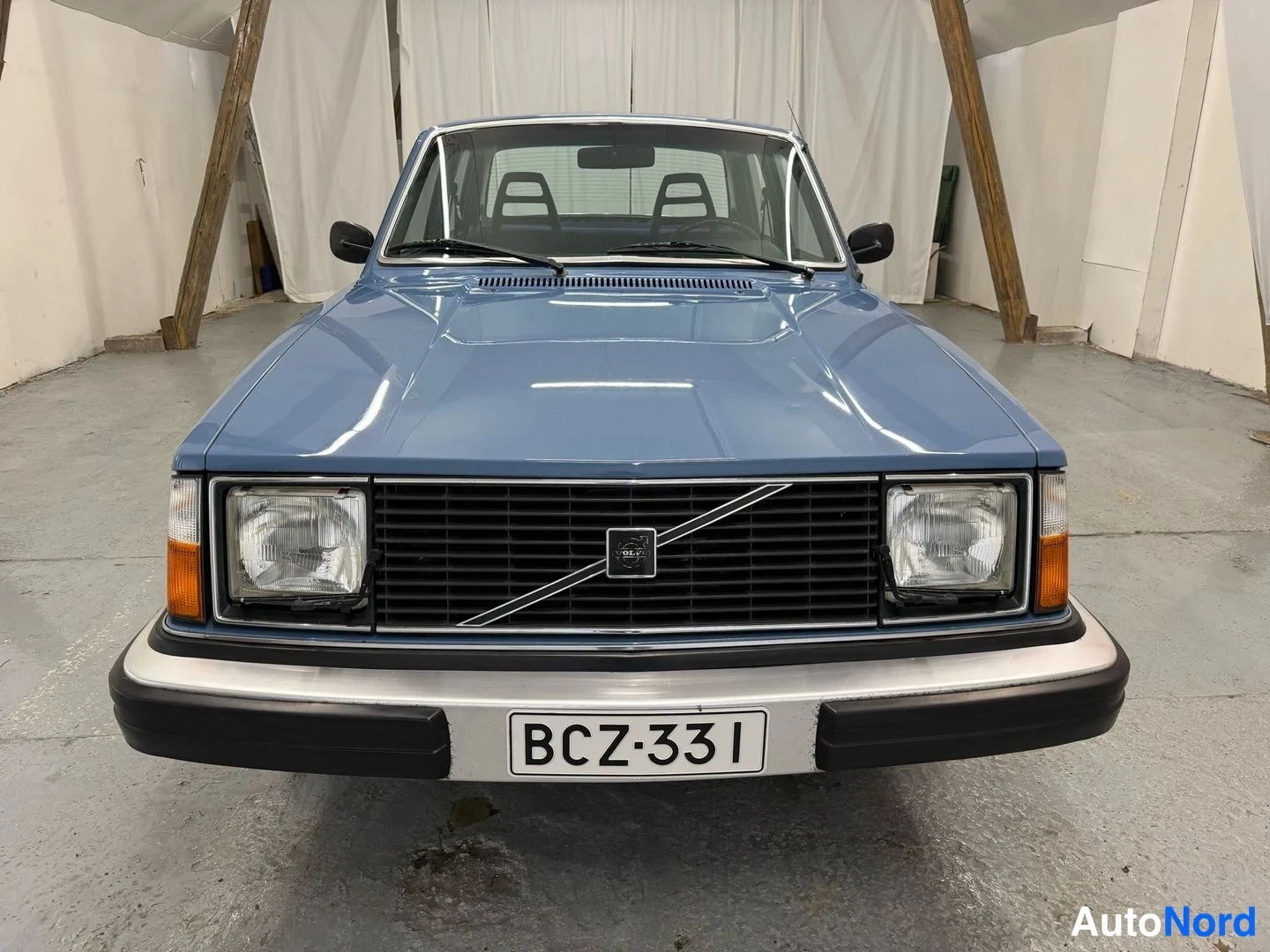 Edestä: Sinisen Volvo 240 -sedan keula suoraan edestä; musta etusäleikkö Volvo-merkillä, pyöreät ajovalot ja rekisterikilpi.
