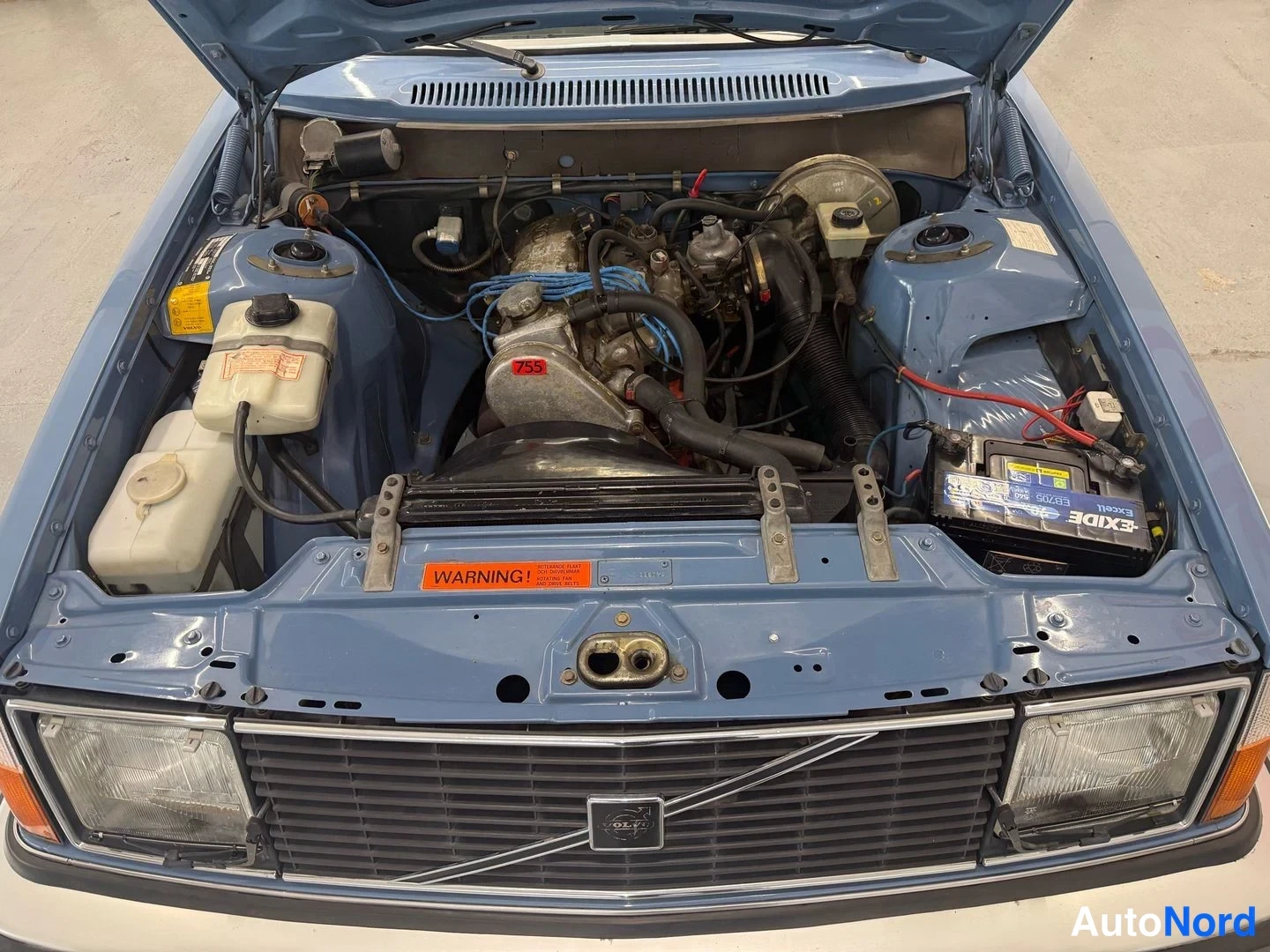 Edestä: Avattu moottoritila sinisessä Volvo 240:ssä, josta näkyvät moottori, Exide-akkupletera, jäähdytinjärjestelmän paisuntasäiliöt ja ämpärin kansi