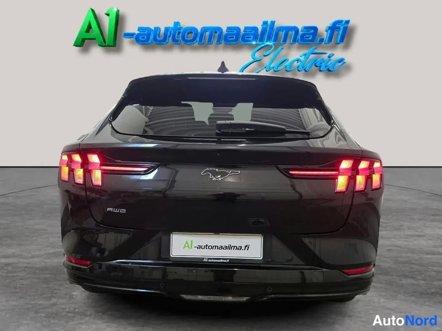 Takaa: Mustan crossoverin takaosa suoraan takaa: hehkuvat poikittain asennetut takavalot, Ford-Mustang-merkki takaoven keskellä, AWD-merkki ja myyjän A1-automaa.ma.fi-