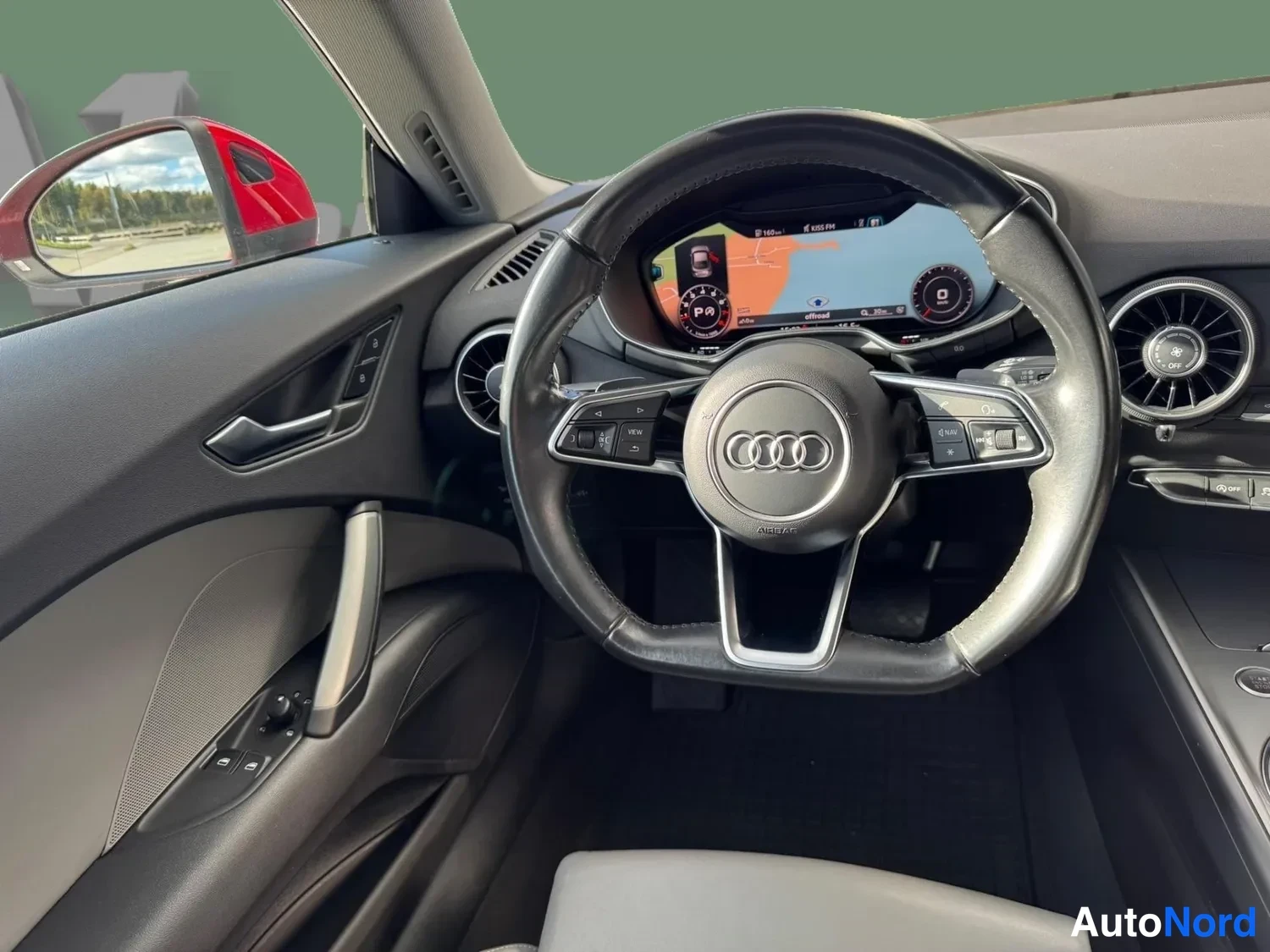 Muu: Audi TT:n ohjaamo: Audi-merkitty monitoimiohjauspyörä, digitaalinen mittaristo ja pyöreät ilmastointisuuttimat, kuljettajan ovenikkuna vasemmalla.