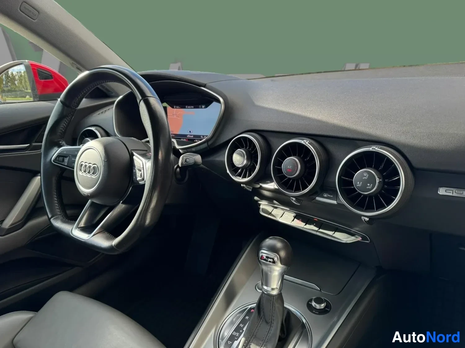 Etuviisto: Ohjaamo näkyvissä: Audi-ohjauspyörä, digitaalinen mittaristonäyttö kartalla, kolme pyöreää ilmastointisuutinta ja automaattivaihteen valitsin.