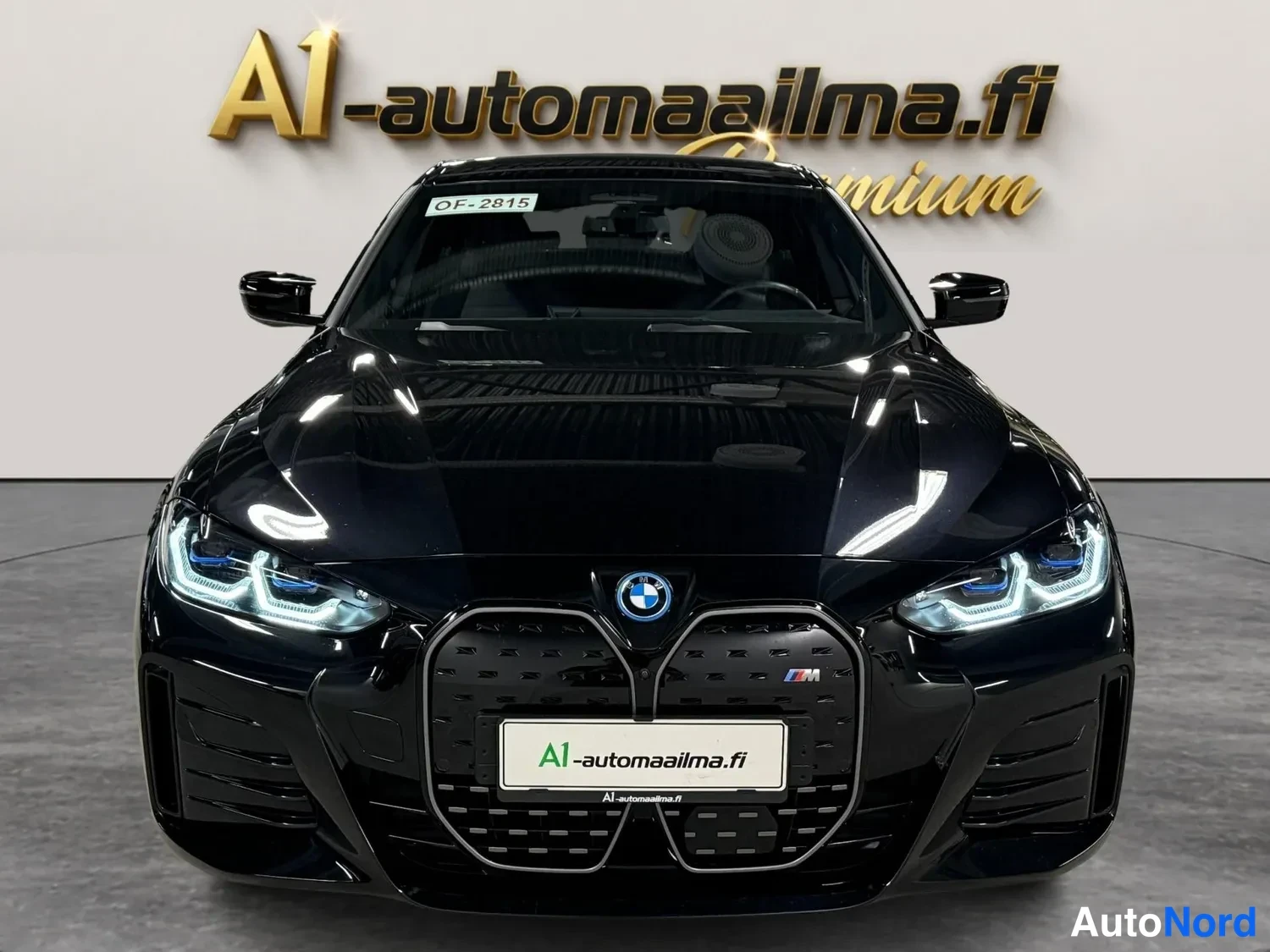 Edestä: Mustan BMW i4:n suora etunäyttö: LED-ajovalot, BMW-merkki ja M-tunnuksella varustettu suljettu etusäleikkö.