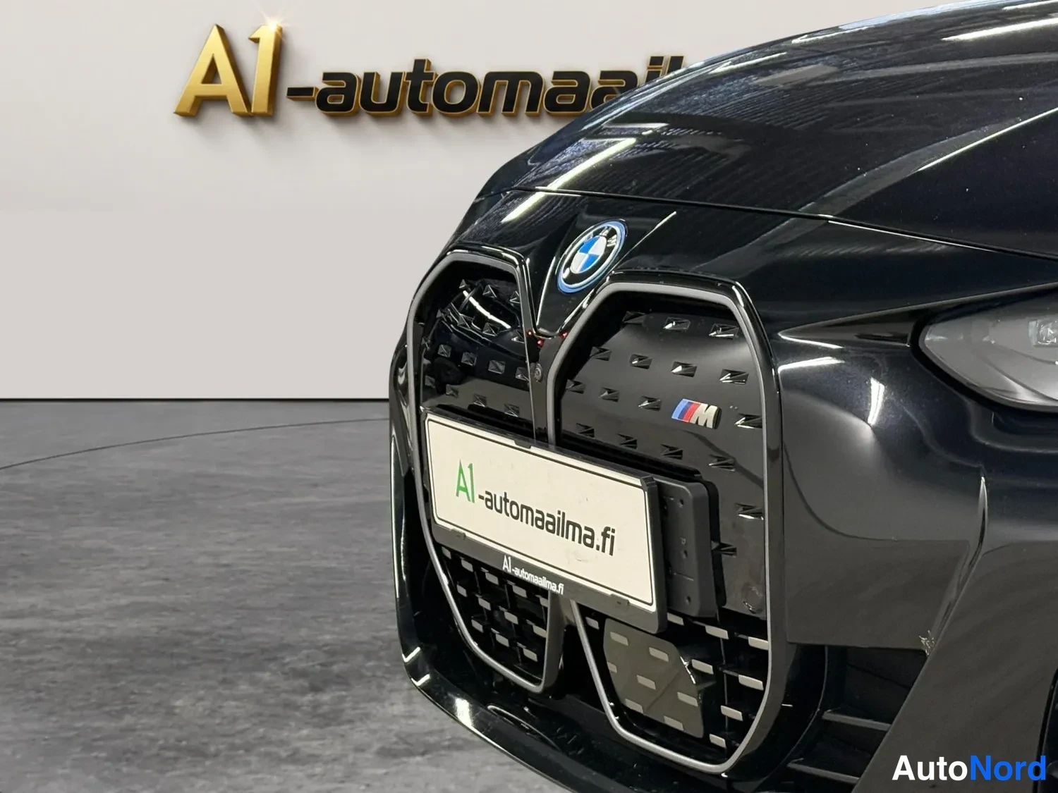 Edestä: Mustan auton etunosa lähikuvassa: BMW-merkki konepellin nokassa, suuret suljetut etusäleiköt ja M-merkki. Puskurissa A1-automaailma-tunnus. Taustalla valkoinen
