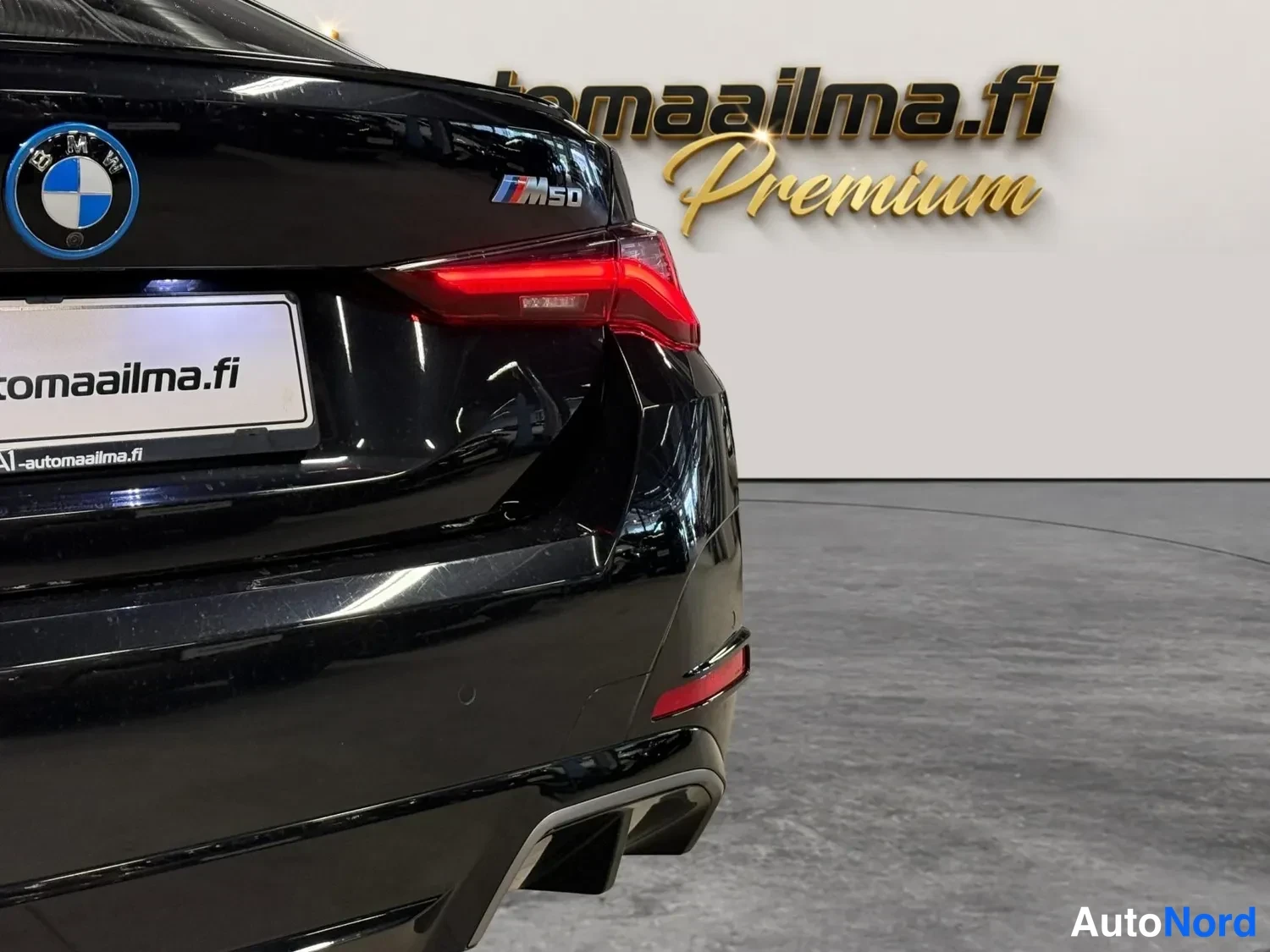 Takaa: Mustan auton takaosa: BMW-emblemi, M50-merkki ja takavalo; rekisterikilpi peitetty mainoskilvellä.