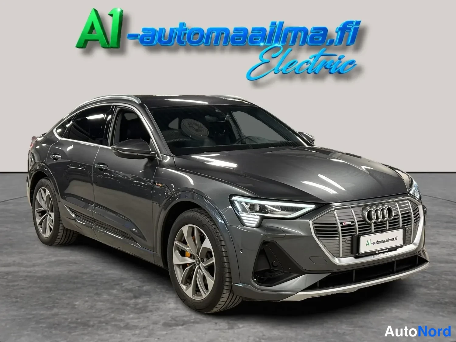 Etuviisto, nokka oikealle: Tummanharmaa Audi e-tron Sportback -tyyppinen sähköauto kulmassa edestä vasemmalta kuvattuna myyntihallissa.