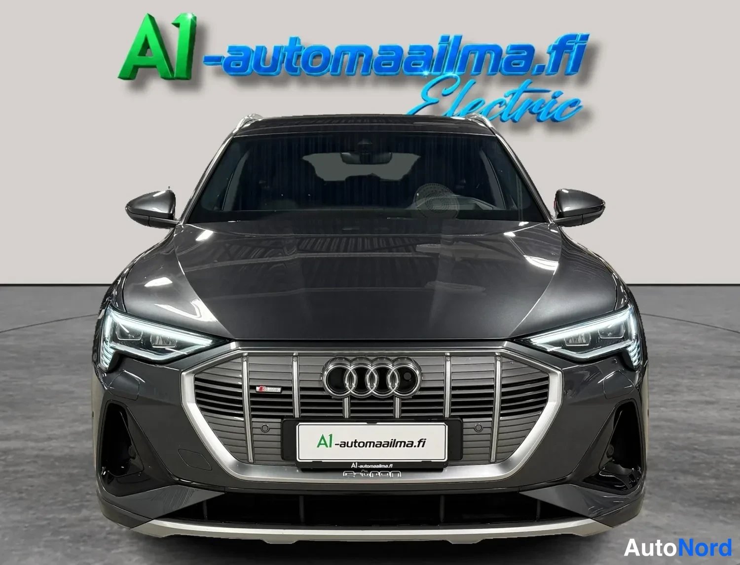 Edestä: Tummanharmaan Audi e-tron -auton etupää suoraan edestä kuvattuna, säleikössä Audi-logot ja A1-automailma-kyltti.