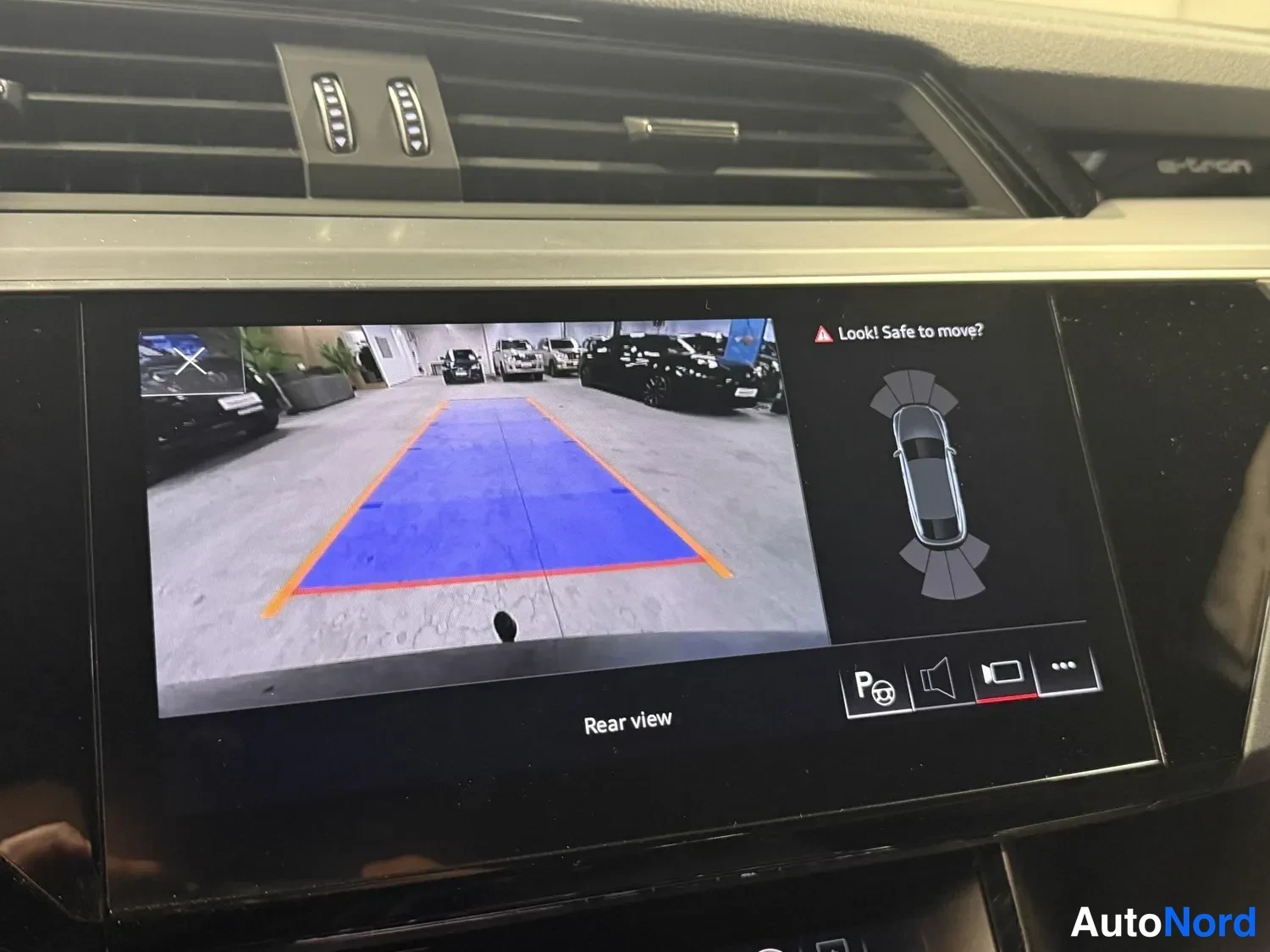 Muu: Audi e-tronin keskinäytön taaksepäin kääntyvä kamera -näkymä (Rear view) sinisellä oppaansuuntausalueella, paneelin viereinen teksti Look! Safe to move? ja auto