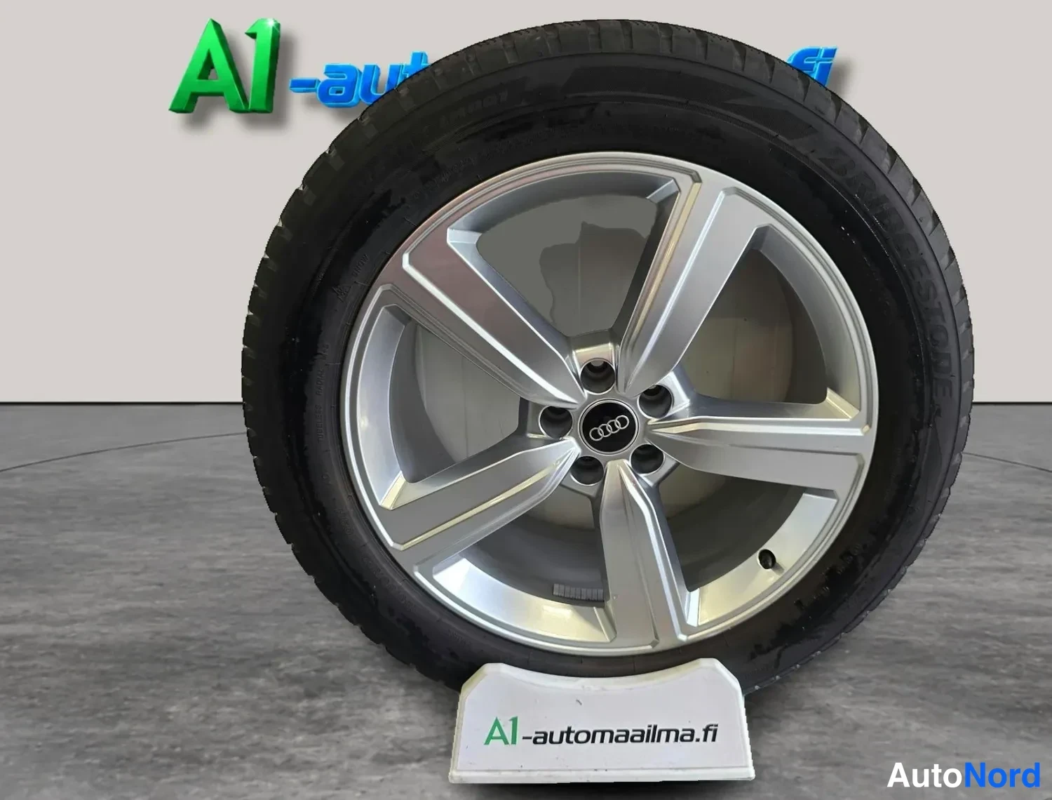 Yksityiskohta: Hopeinen Audi-merkkinen viisisakarainen kevytmetallivanne, musta rengas, taustalla A1-automaailma.fi-logo.