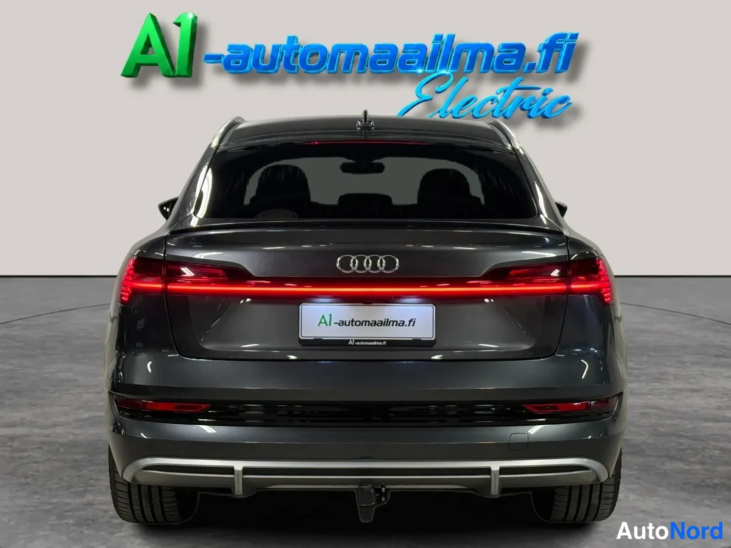 Takaa: Takanäkymä tummanharmaasta autosta, valaistut poikittaiset takavalot, takaikkuna ja vetokoukku.