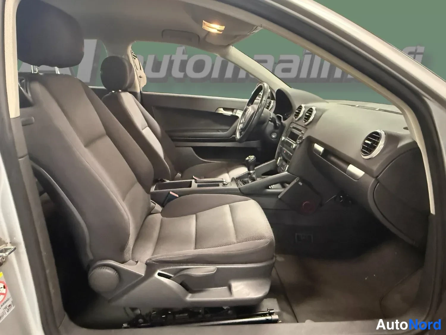 Muu: Audi A3:n matkustamo toiselta puolelta: kaksi etuistuinta, ohjauspyörä ja kojelauta näkyvät avoimesta ovenasta.