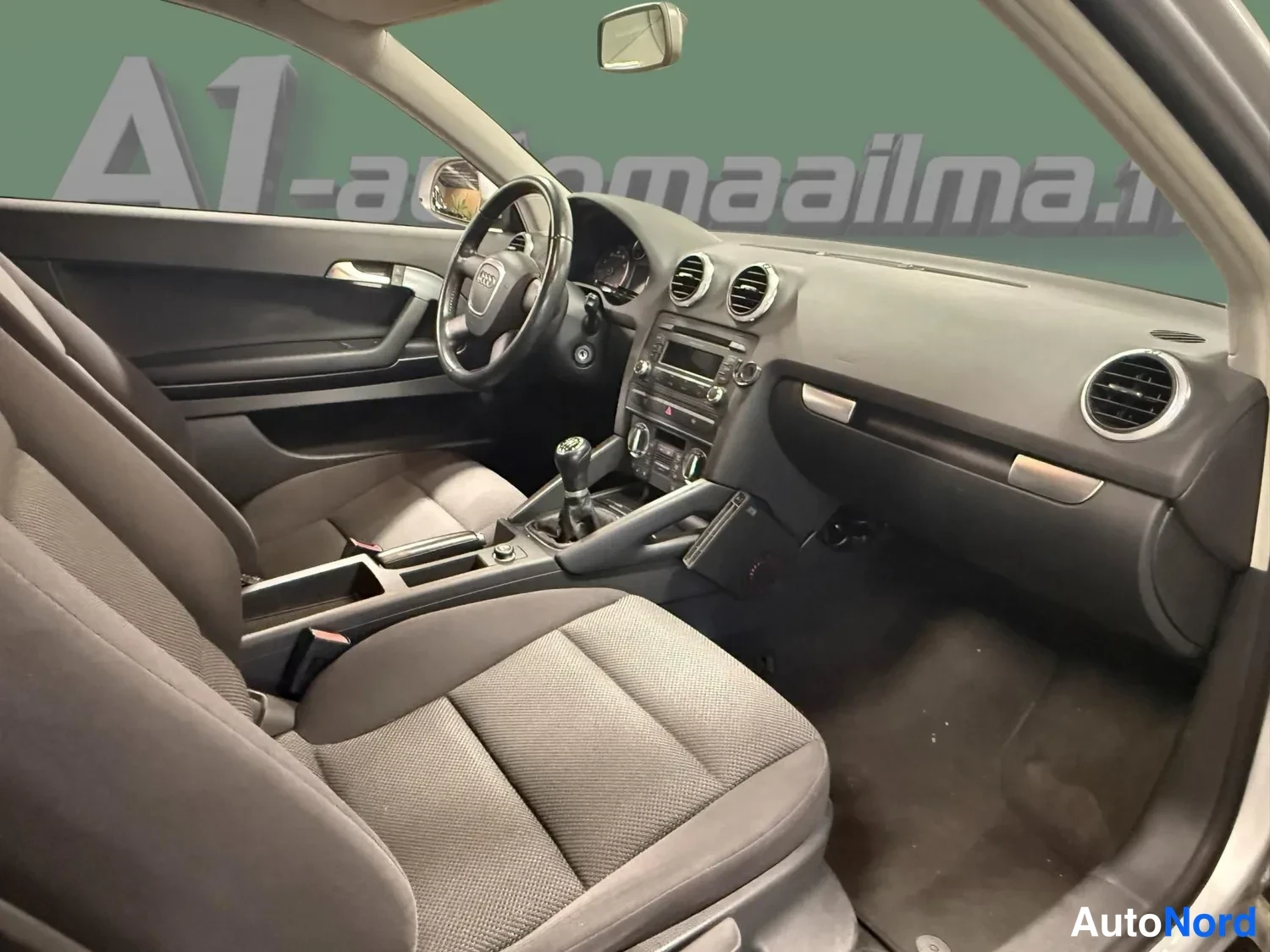 Muu: Audi A3:n kojelauta ja matkustamo: Audi-ohjauspyörä, keskikonsoli, ilmastointiritilät ja tummat kangasistuimet näkyvät.