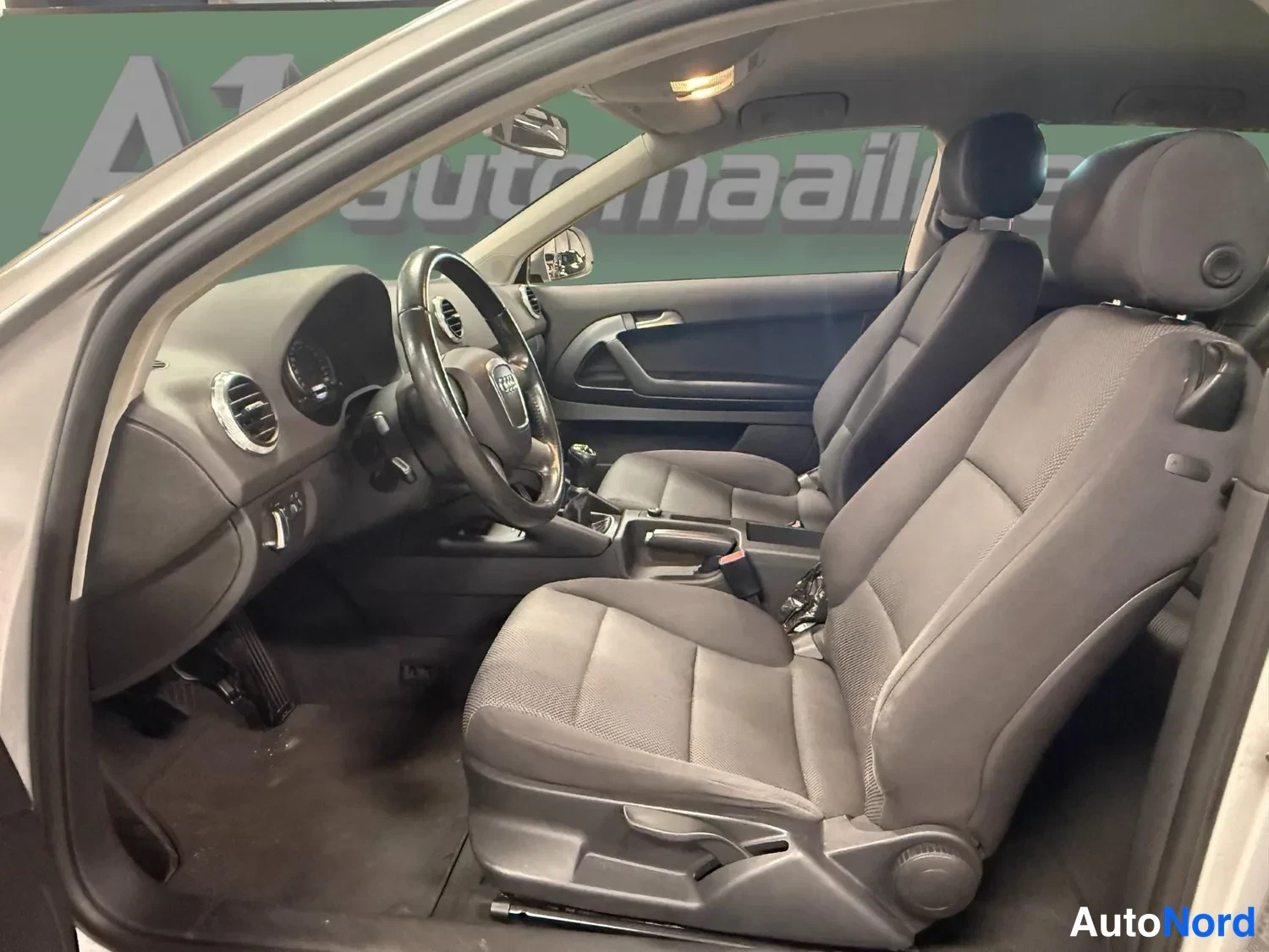 Muu: Audi A3:n matkustamo sivulta: tummat kankaanpenkit, ohjauspyörä ja kojelauta näkyvät avoimesta ovenasta.