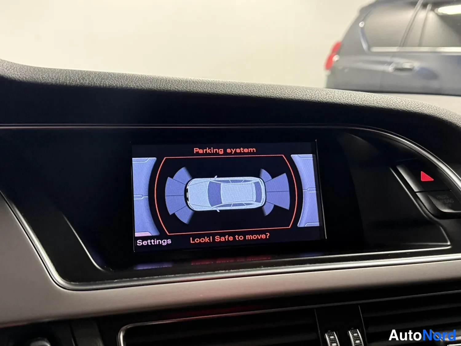 Yksityiskohta: Kojetaulun näyttö näyttää pysäköintijärjestelmän (Parking system) auton ympäriltä, jossa on anturipalkit, sekä tekstin 'Look! Safe to move?' ja 'Settings'.