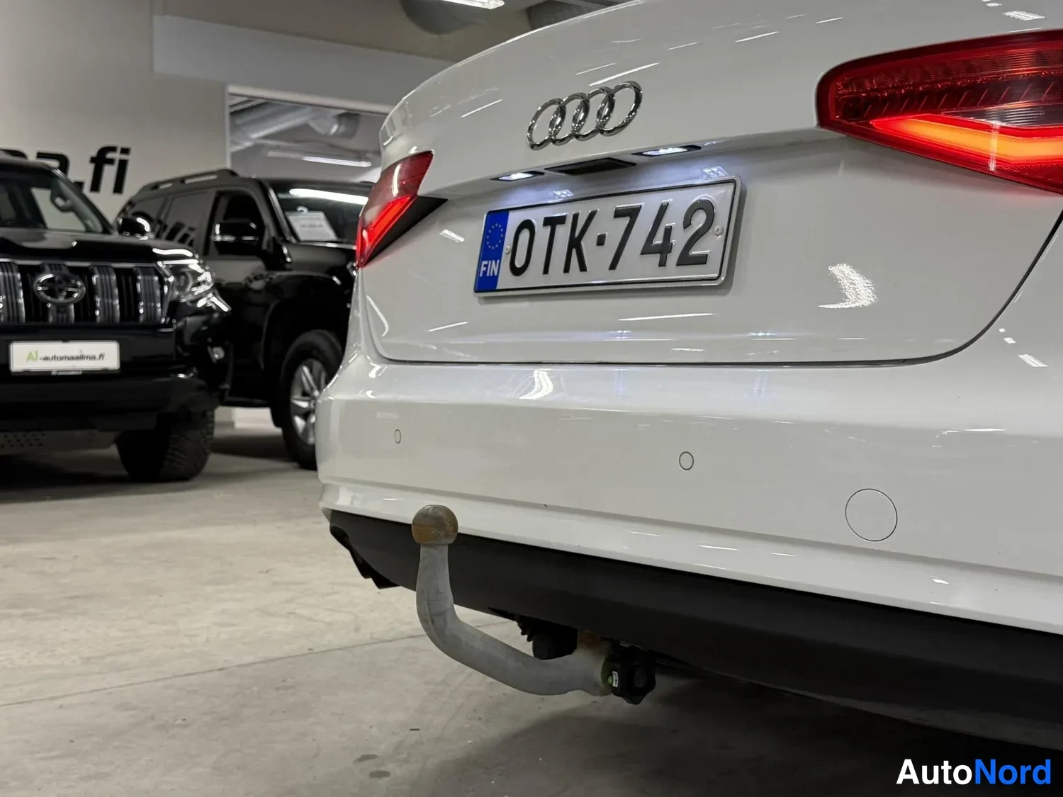 Takaa: Valkoisen auton takapään lähikuva: Audi-merkki, punainen rekisterikilpi FIN ja kiinteä vetokoulu.