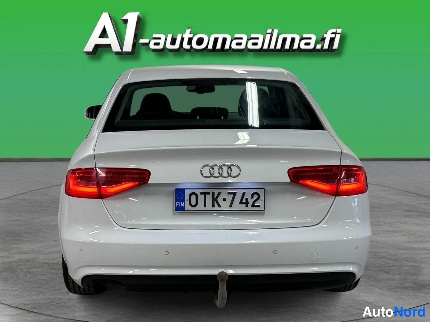 Takaa: Auton takaosa suoraan takaa: valkoinen sedan, punaiset takavalot, Audi-merkit, rekisterikilpi ja vetoaiman pallo takapuskurin alla.