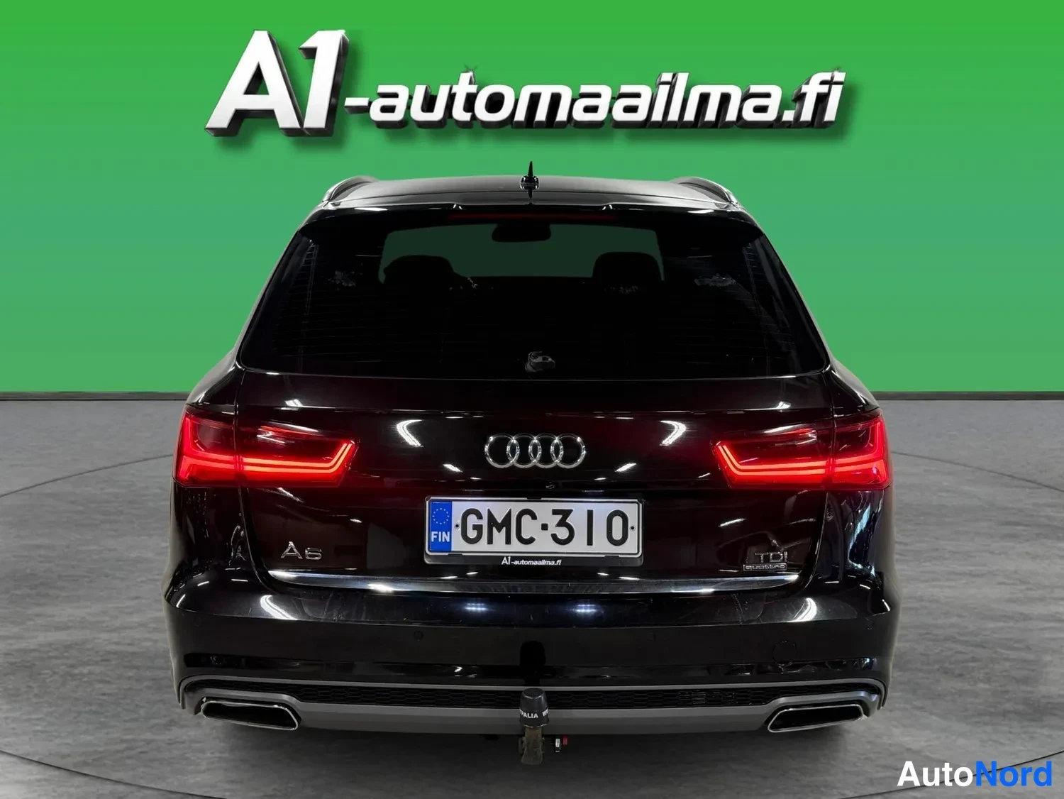 Takaa: Mustan farmariauton takaosa: Audi-merkki, A6-merkki, rekisteritunnus, punaiset takavalot, kaksinkertaiset pakoputket ja vetokoukku.