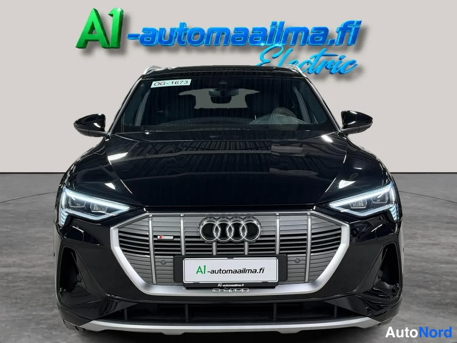 Edestä: Mustan Audi e-tronin suora etukuva, jossa näkyvät pystysäleinen etusäleikkö Audi-embleemilla, LED-ajovalot ja tuulilasiin kiinnitetty OG-1673-tunniste.