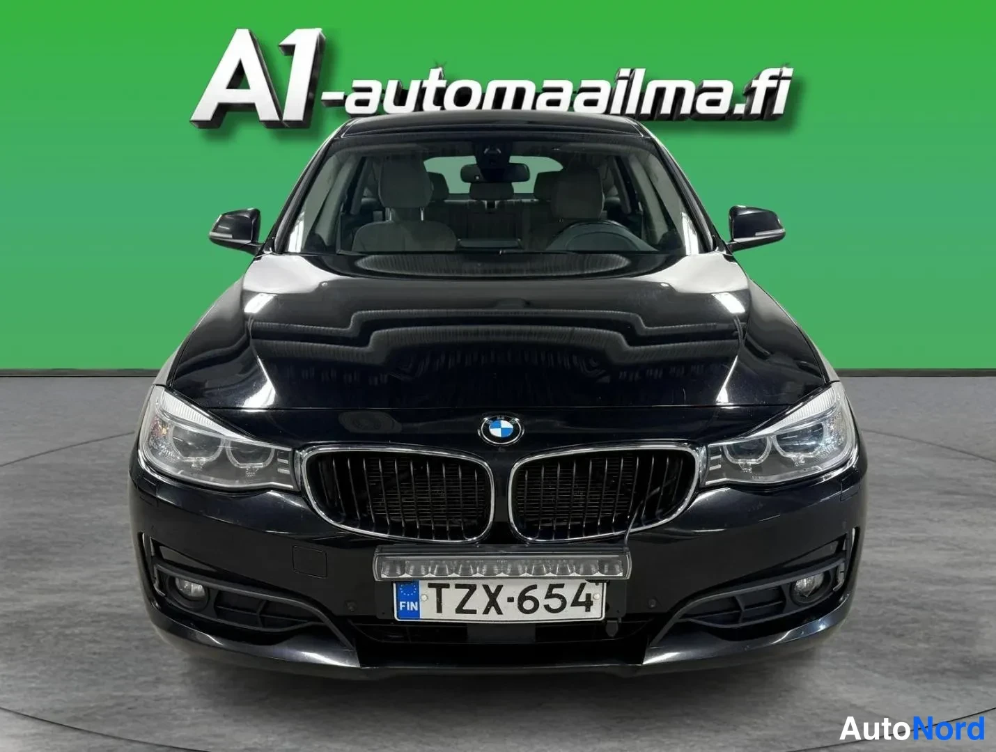 Edestä: Mustan BMW:n suora etukuva, jossa näkyvät etusäleikkö, ajovalot ja etupuskurin rekisterikilpi vihreää taustaa vasten.