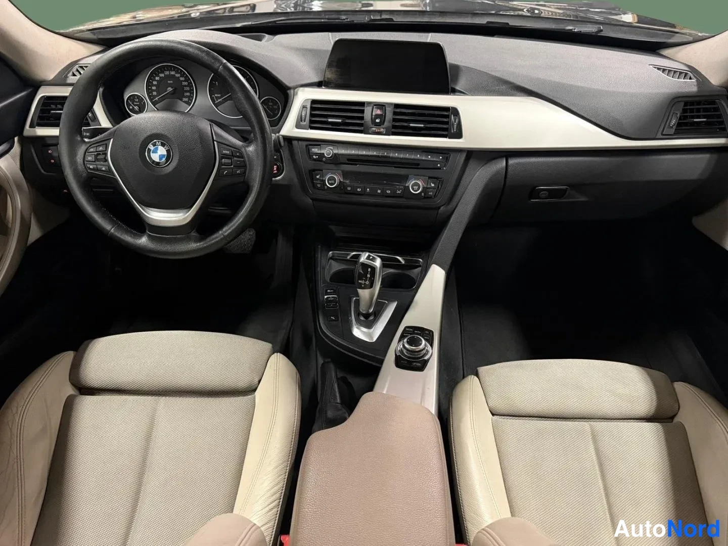 Sivulta: Autojen sisätilakuva keskikäytävän yli: BMW:n ohjaamo, beige-punaiset istuimet, BMW-merkki ohjauspyörässä, automaattivaihteiston valitsin ja iDrive-ohjain.