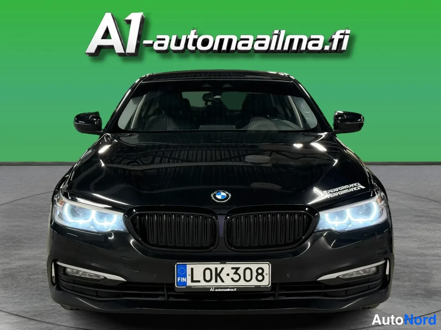Edestä: Musta sedan suoraan edestä, LED-etuvalot päällä, rekisteri.