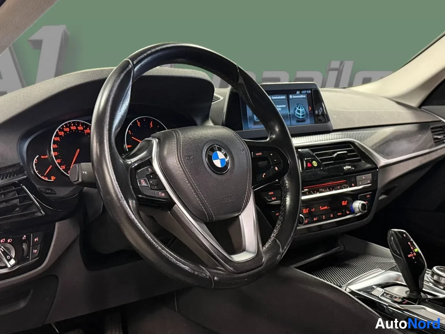 Edestä: Ohjaamo näkyvissä: BMW-ohjauspyörä, analogiset ja digitaaliset mittarit, keskellä näyttö ja automaattivaihteen valitsin.