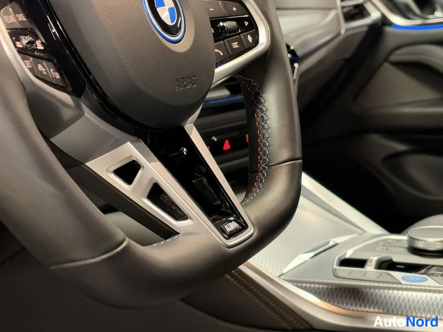 Muu: BMW-ohjauspyörän keski-alaosaa läheltä kuvattuna, AIRBAG-teksti, punaiset ompeleet ja keskikonsolin kytkimiä.