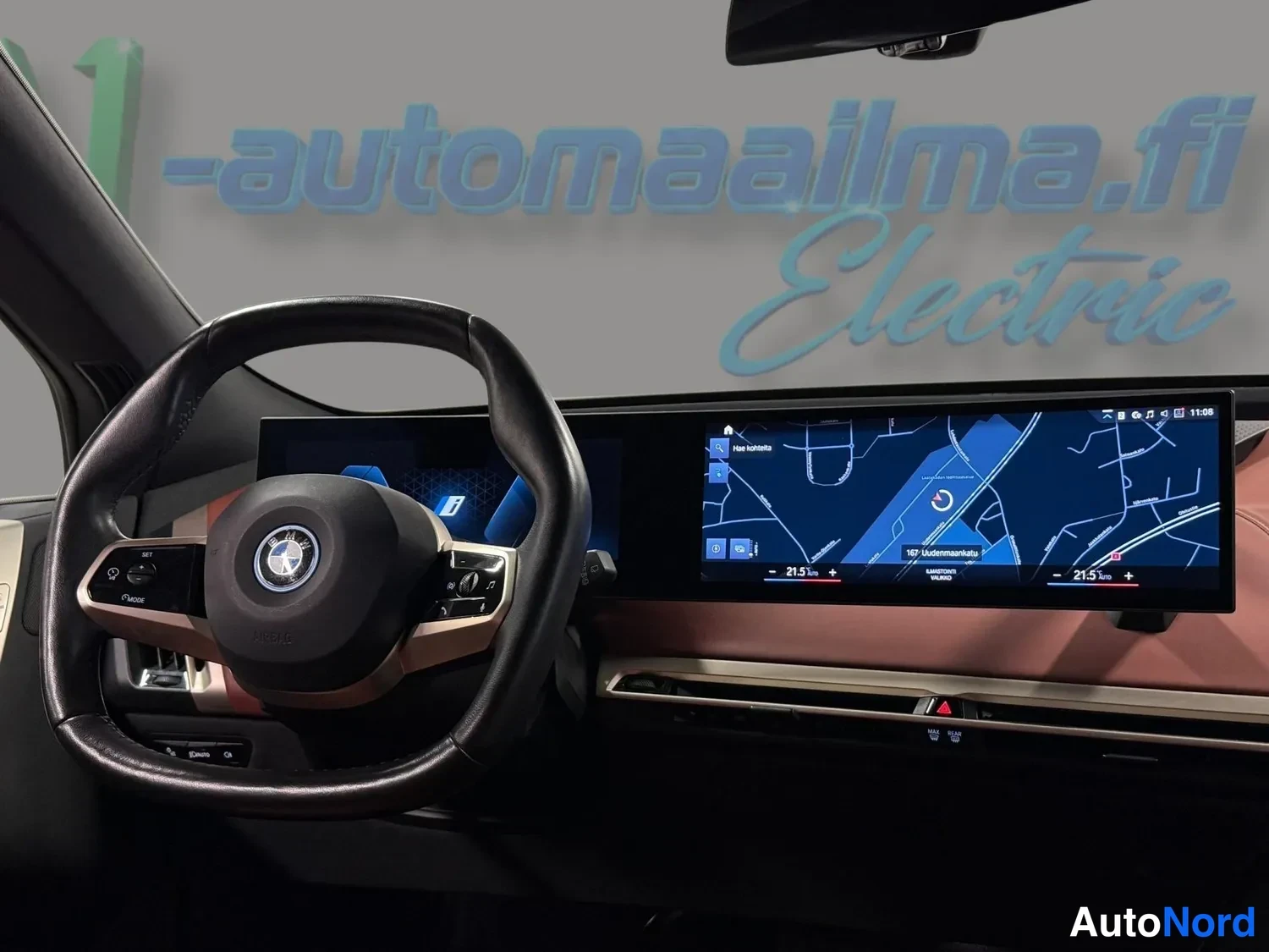 Sivulta: Kuljettajan näkökulma: BMW-ohjauspyörä ja leveä kaareva näyttö, jolla näkyy kartanavigaatio ja kulmatiedot (21,5 kWh/100 km).