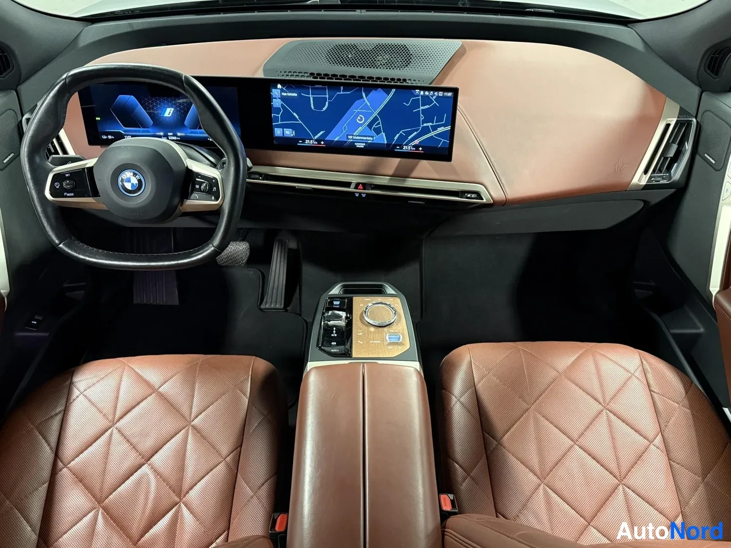 Sivulta: Kojelauta ylhäältä: BMW-ohjauspyörä, kaareva infotainment-näyttö karttaan, ruskea nahkapanelointi ja timanttikuvioiset etuistuimet.