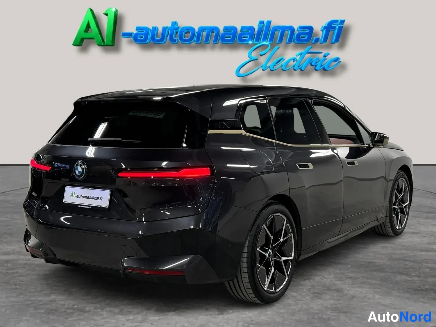 Takaviisto: Tummanharmaa BMW-katemaasturi takaa-vinovasemmalta kuvattuna autosalissa, mustat vanteet ja pitkulaiset punaiset takavalot.