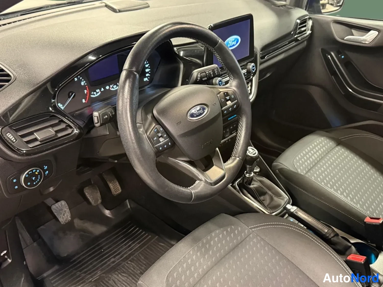 Muu: Ford Fiesta ohjaamo edestä: ohjauspyörä Ford-merkillä, kojelauta, kosketusnäyttö, manuaalivaihtoheitin ja tummat kangasistuimet.