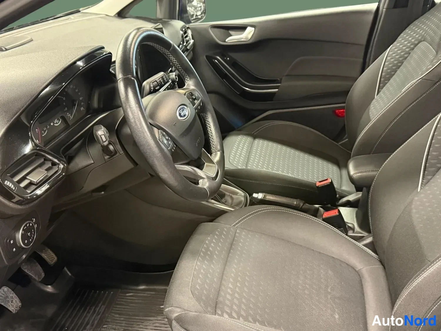 Muu: Ford Fiesta ohjaamon sisäkuva: ohjauspyörä, kojelauta, matkustajan puolen ovi ja tummat kangasistuimet kuvattuna oikealta.