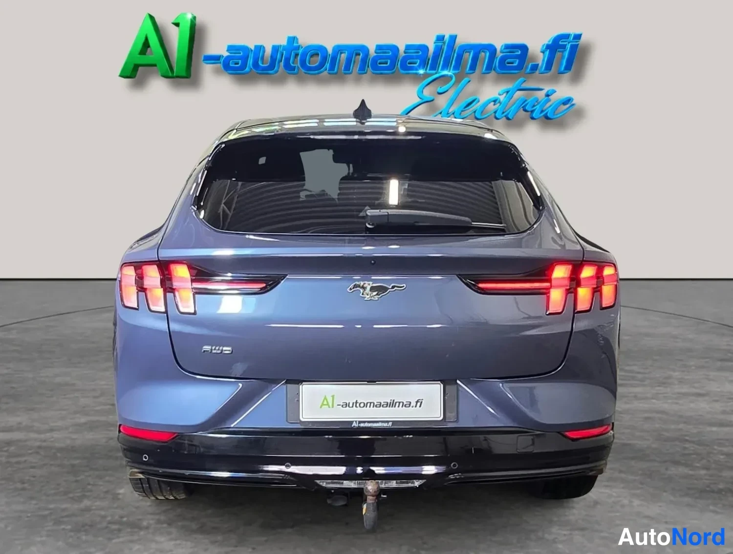 Takaa: Sinertävän harmaa Ford Mustang Mach-E suoraan takaa valaistuine takavaloina, kylkipeilillä ja perävaunukoukulla.